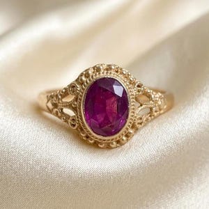 May include: A gold ring featuring an oval-cut deep pink gemstone set in a decorative metal frame with intricate filigree detailing and a beaded border. The band is thin and smooth, connecting to the ornate setting that holds the central stone.