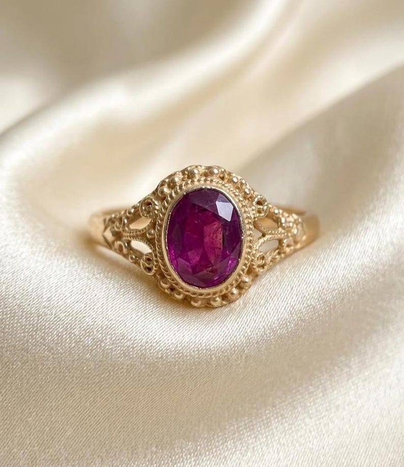May include: A gold ring featuring an oval-cut deep pink gemstone set in a decorative metal frame with intricate filigree detailing and a beaded border. The band is thin and smooth, connecting to the ornate setting that holds the central stone.