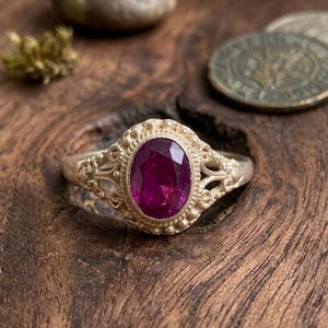 May include: A silver ring featuring an oval-cut, deep magenta gemstone set in a decorative metal frame. The band and setting display intricate filigree metalwork with small beaded accents surrounding the central stone, creating a detailed texture.
