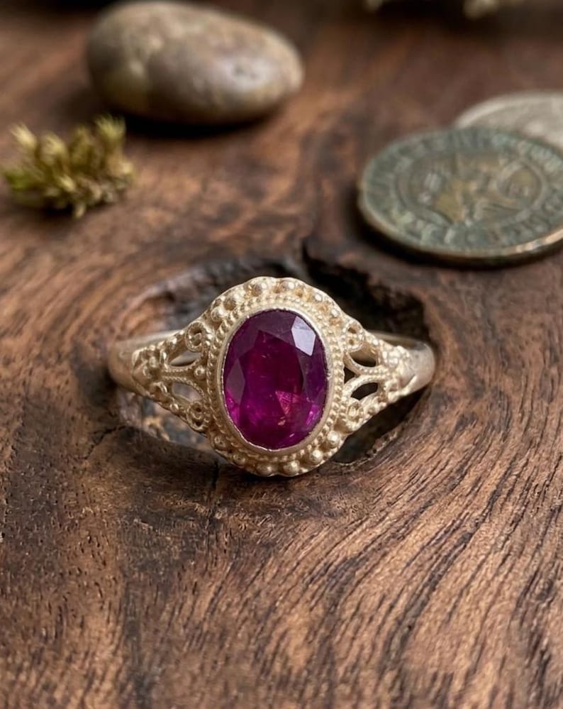May include: A silver ring featuring an oval-cut, deep magenta gemstone set in a decorative metal frame. The band and setting display intricate filigree metalwork with small beaded accents surrounding the central stone, creating a detailed texture.