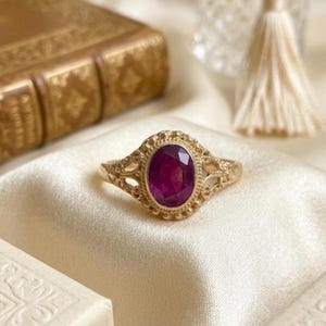 May include: A gold ring featuring an oval-cut deep pink gemstone set in a decorative metal frame with intricate filigree detailing along the band. The setting is surrounded by a beaded border, creating a textured appearance around the central stone.