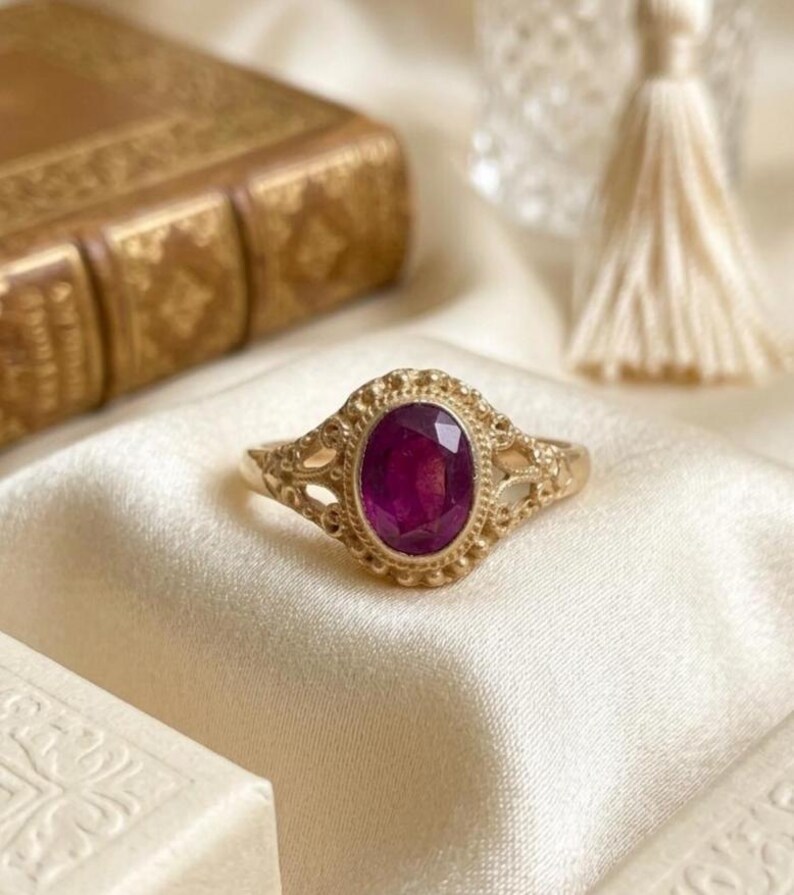 May include: A gold ring featuring an oval-cut deep pink gemstone set in a decorative metal frame with intricate filigree detailing along the band. The setting is surrounded by a beaded border, creating a textured appearance around the central stone.