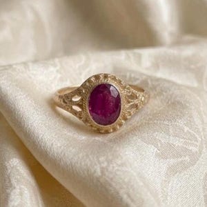 May include: A gold ring featuring an oval-shaped, deep pinkish-red gemstone set in the center. The metal band displays intricate, textured filigree detailing around the stone setting, creating a decorative border with small, raised bead-like accents.