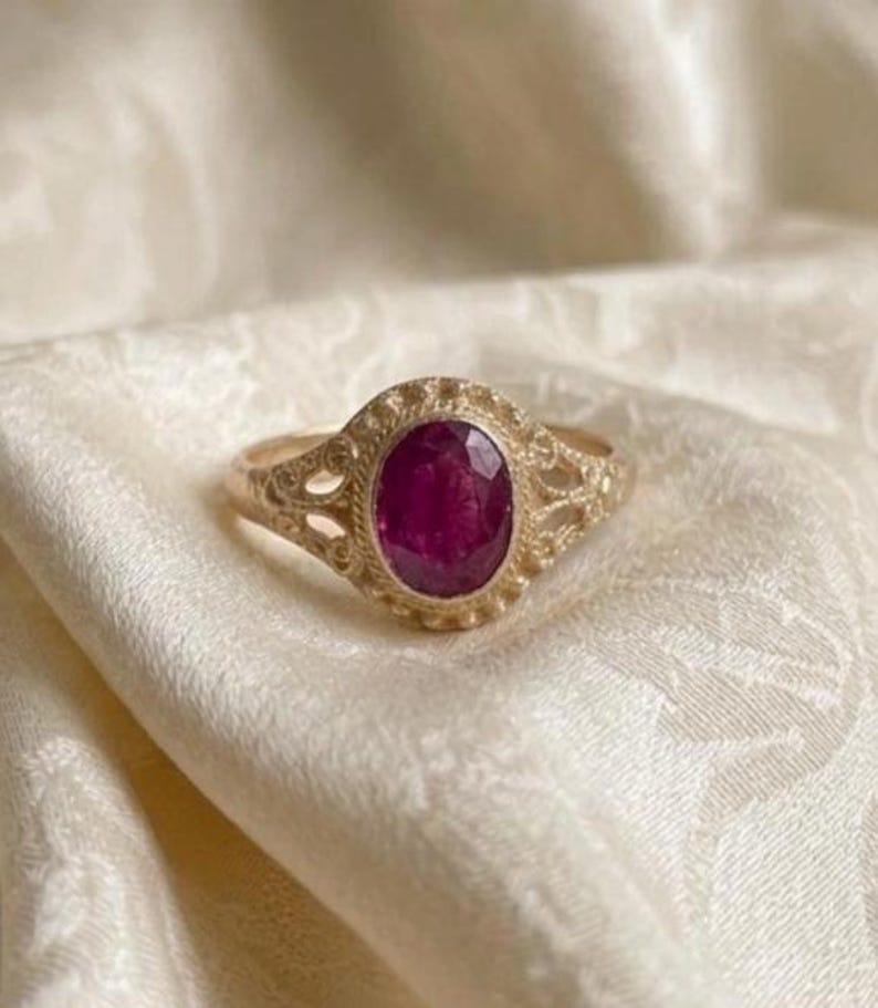 May include: A gold ring featuring an oval-shaped, deep pinkish-red gemstone set in the center. The metal band displays intricate, textured filigree detailing around the stone setting, creating a decorative border with small, raised bead-like accents.