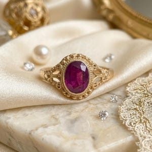 May include: A gold ring featuring an oval-cut deep pink gemstone set in a decorative filigree band with beaded metal detailing around the stone. The ring has an intricate openwork design on the sides of the band, showcasing a polished finish.