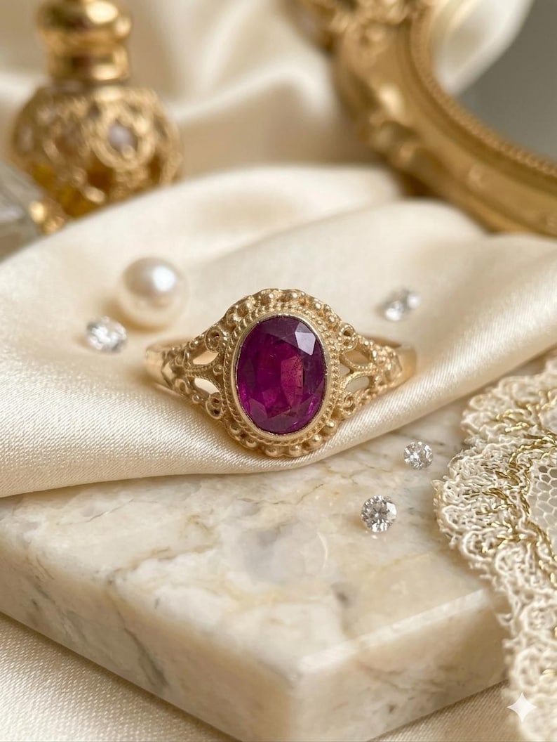 May include: A gold ring featuring an oval-cut deep pink gemstone set in a decorative filigree band with beaded metal detailing around the stone. The ring has an intricate openwork design on the sides of the band, showcasing a polished finish.