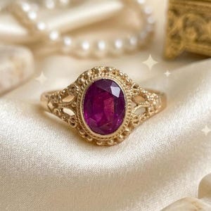 May include: A gold ring featuring an oval-cut, deep magenta-colored gemstone set in a decorative, beaded metal frame. The band displays intricate, openwork metal detailing on the sides, creating a delicate and ornate appearance for this jewelry piece.
