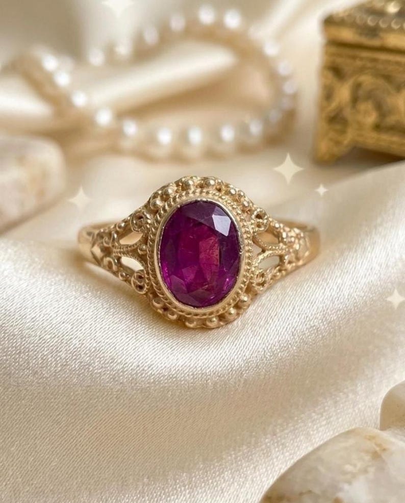 May include: A gold ring featuring an oval-cut, deep magenta-colored gemstone set in a decorative, beaded metal frame. The band displays intricate, openwork metal detailing on the sides, creating a delicate and ornate appearance for this jewelry piece.