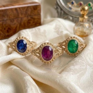 May include: Three gold rings featuring intricate filigree bands and oval-cut gemstones. The left ring has a deep blue sapphire, the center ring holds a vibrant magenta ruby, and the right ring displays a brilliant green emerald gemstone.