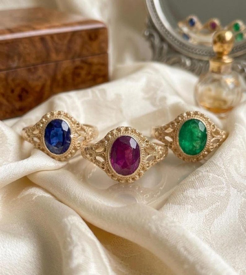 May include: Three gold rings featuring intricate filigree bands and oval-cut gemstones. The left ring has a deep blue sapphire, the center ring holds a vibrant magenta ruby, and the right ring displays a brilliant green emerald gemstone.