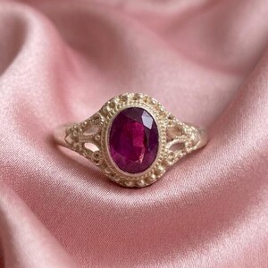 May include: A silver ring featuring an oval-cut deep pink gemstone set in a decorative metal frame with beaded detailing and intricate openwork patterns along the band, showcasing a polished finish and a prominent central stone setting.