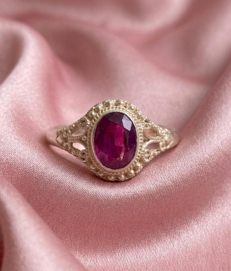 May include: A silver ring featuring an oval-cut deep pink gemstone set in a decorative metal frame with beaded detailing and intricate openwork patterns along the band, showcasing a polished finish and a prominent central stone setting.