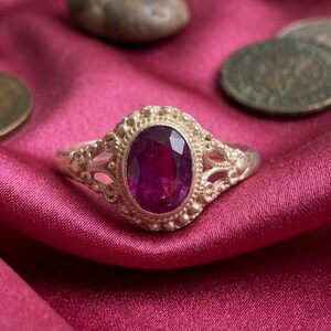 May include: A silver ring featuring an oval-cut deep pink gemstone set in a decorative bezel. The band has an intricate openwork design with small metal beads surrounding the central stone, creating a textured appearance around the gemstone.