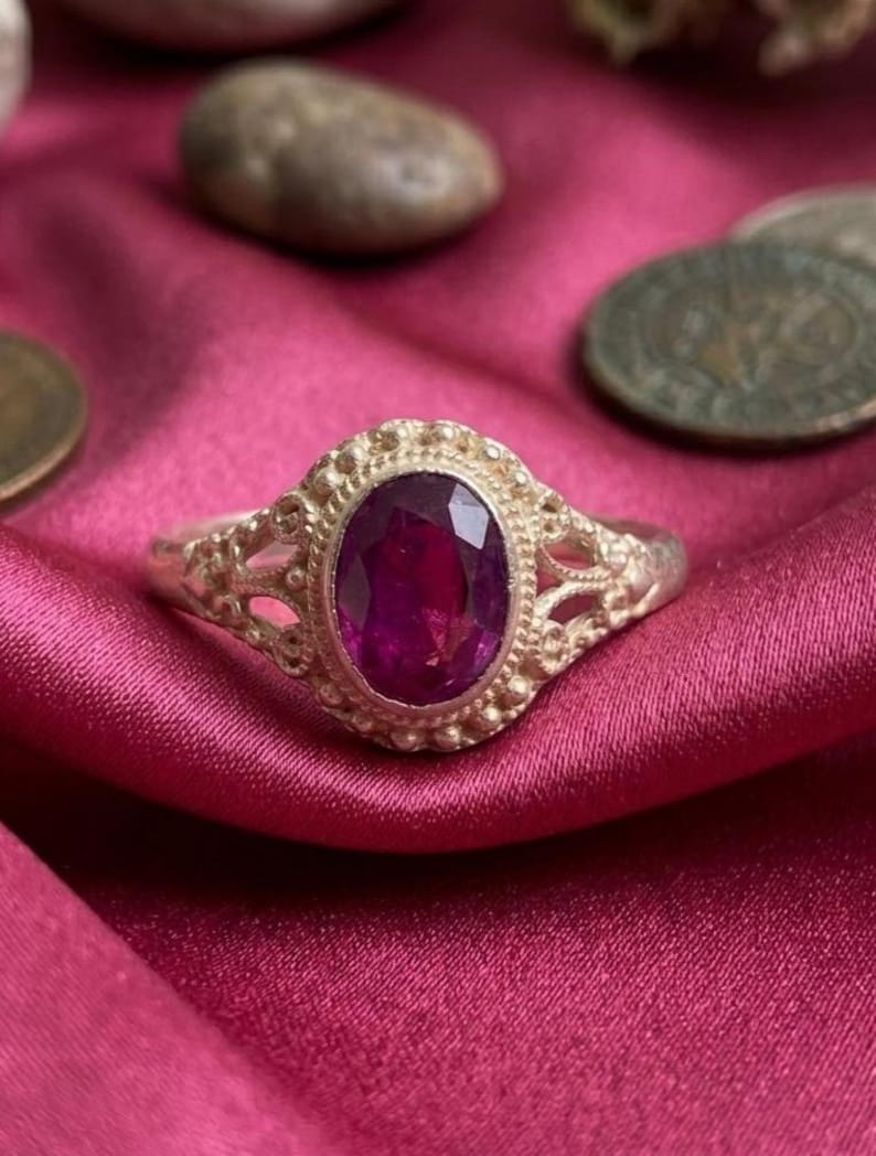 May include: A silver ring featuring an oval-cut deep pink gemstone set in a decorative bezel. The band has an intricate openwork design with small metal beads surrounding the central stone, creating a textured appearance around the gemstone.