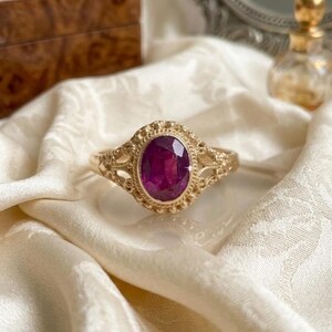 May include: A gold ring featuring an oval-cut, deep magenta gemstone set in a decorative, textured metal frame. The band displays intricate, openwork filigree detailing on both sides, surrounding the central stone with a beaded metal border.