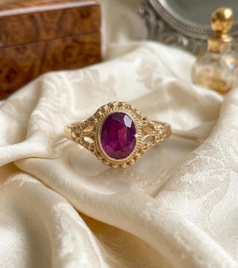May include: A gold ring featuring an oval-cut, deep magenta gemstone set in a decorative, textured metal frame. The band displays intricate, openwork filigree detailing on both sides, surrounding the central stone with a beaded metal border.