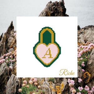 Könnte beinhalten: Ein Kreuzstich-Design eines herzförmigen Schlosses mit dem Buchstaben "A" darin. Das Schloss ist grün und gold umrandet, mit einem rosa Herz in der Mitte. Das Wort "Riche" steht darunter in Kursivschrift.