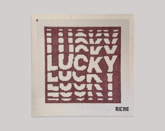 Op de afbeelding: Een vierkant naaldwerk canvas met een donkerrood blokontwerp waarin het woord LUCKY wordt herhaald in een vervormd, golvend lettertype. Het canvas heeft een witte rand en het woord RICHE is in kleine zwarte letters in de rechterbenedenhoek geborduurd.