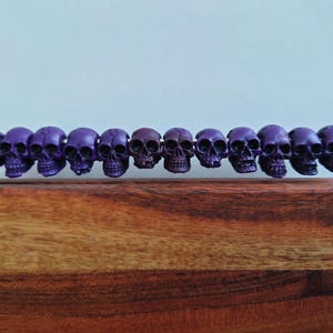 May include: A row of eleven purple skull beads strung on a silver metal rod. The skulls are detailed with eye sockets, noses, and teeth. The skulls are displayed on a wooden surface with a light blue background.