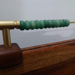 Può includere: Un oggetto decorativo in stile abaco in legno e metallo. La base è un quadrato marrone scuro, che supporta un palo di metallo color oro e una barra orizzontale. La barra contiene una serie di perle verdi e arrotondate. L'abaco è su uno sfondo chiaro.