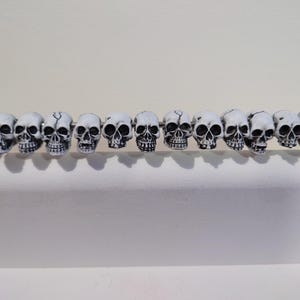 May include: Eleven small, white skull beads with black detailing strung on a silver rod. The skulls have a cracked appearance and are attached to a wooden handle on the left. A decorative item featuring skull beads.
