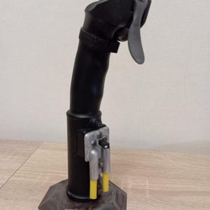 May include: A black joystick with a grey base. The joystick has a curved handle with a trigger and a button. The base is a multi-sided polygon shape. Two yellow tubes are visible near the base.