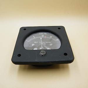 May include: A black voltmeter with a square frame and a circular gauge. The gauge reads "VOLTS" and displays a needle pointing to 10. The frame has four screw holes and a central screw. The text "REF. N 5A/1630 A/M" is visible.