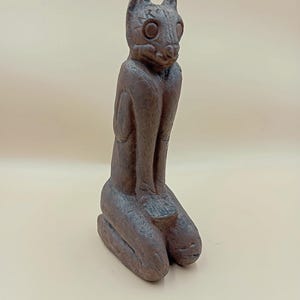 Peut inclure: Statue de chat marron agenouillé. Le chat a de grands yeux ronds et des oreilles pointues. La statue est faite d'un matériau texturé et a un aspect rustique. Le chat est agenouillé, les pattes jointes.