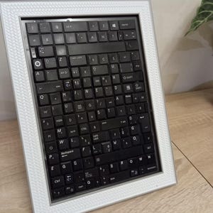 Può includere: Un'opera d'arte incorniciata con una tastiera di computer nera. La tastiera è inserita in una cornice bianca con un bordo decorativo strutturato. I tasti, con lettere e simboli, sono visibili. L'opera è esposta su una superficie marrone chiaro.
