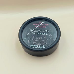 Op de afbeelding: Een zwarte brandstofmeter met witte tekst en een naald die het brandstofpeil aangeeft. De meter geeft "GALLONS FUEL" en "NORMAL FLIGHT" aan. De meter heeft een capaciteit van 182 liter.