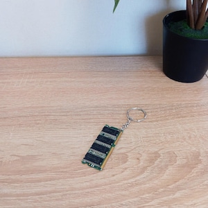 May include: A computer RAM module keyring with a silver ring. The RAM module is green and black with gold connectors. The keyring is on a light brown wooden surface.