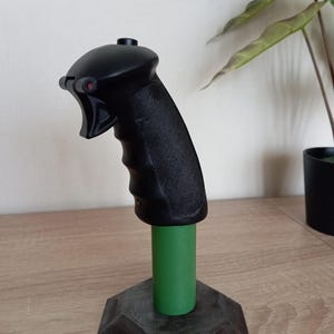May include: A black joystick with a textured grip, a green cylindrical base, and a gray, multi-sided base. The joystick has a red button and is designed for gaming or flight simulation.