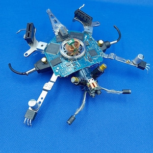 Könnte beinhalten: Eine einzigartige, abstrakte Skulptur aus recycelten elektronischen Bauteilen. Das Kunstwerk zeigt eine zentrale blaue Leiterplatte mit verschiedenen angebrachten Teilen, darunter Metallhalterungen, Drähte und Anschlüsse, die in einem radialen Muster vor blauem Hintergrund angeordnet sind.