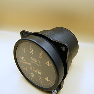 May include: A black aircraft climb and descent indicator with a circular gauge. The gauge has white numerals and markings against a dark background. The text "CLIMB THOUSANDS" and "DESCENT" are visible.