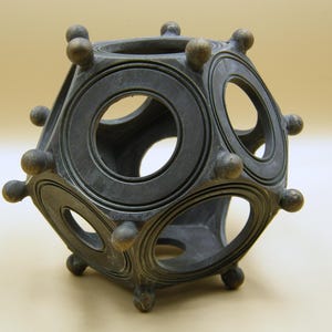 Op de afbeelding: Een donkergrijze, geometrische sculptuur met cirkelvormige en ovale openingen. De sculptuur heeft een complex, veelzijdig ontwerp met afgeronde uitsteeksels op elke hoek. Het stuk lijkt van metaal te zijn en heeft een verweerde uitstraling.