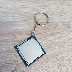 Peut inclure: Un porte-clés CPU argenté avec un porte-clés et une chaîne argentés. Le CPU est de forme carrée avec un dessus argenté et une base vert foncé. Le porte-clés est posé sur une surface en bois brun clair.