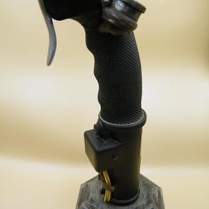 May include: A black joystick with a textured grip and a trigger. The joystick has a cylindrical base and a gray, octagonal stand. Yellow wires are visible near the base. The joystick is designed for use with a flight simulator.