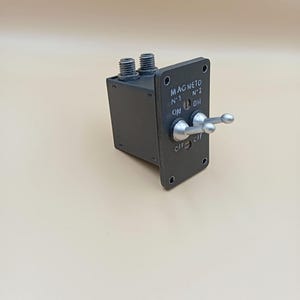 Può includere: Un interruttore magnete grigio scuro con due leve e il testo "MAGNETO N.1 N.2 ON ON OFF OFF". L'interruttore ha due connettori filettati ed è montato su una piastra rettangolare.