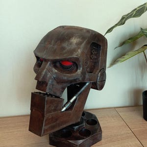May include: A brown, robotic skull sculpture with glowing red eyes. The sculpture has a weathered, metallic finish and is mounted on a hexagonal base with pen holders. A pen is placed in the holder. The sculpture is a unique piece of art.