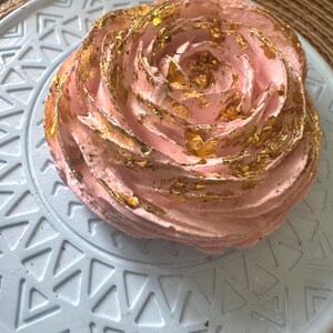 May include: A light pink, rose-shaped meringue dessert topped with delicate gold leaf flakes. The treat is presented on a round, pale blue plate featuring a geometric pattern of embossed triangles and lines, creating a textured surface.