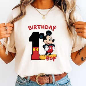May include: Cream-colored t-shirt featuring a graphic of Mickey Mouse with the text "BIRTHDAY 1 BOY" in red and black. Mickey is wearing red shorts and yellow shoes. The number 1 is black with red accents.