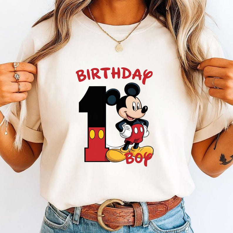 May include: Cream-colored t-shirt featuring a graphic of Mickey Mouse with the text "BIRTHDAY 1 BOY" in red and black. Mickey is wearing red shorts and yellow shoes. The number 1 is black with red accents.