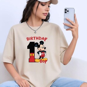May include: Beige t-shirt with a graphic of Mickey Mouse, the number 1, and the words "BIRTHDAY BOY" in red. Mickey is wearing red shorts, black gloves, and yellow shoes. The shirt is a casual style.