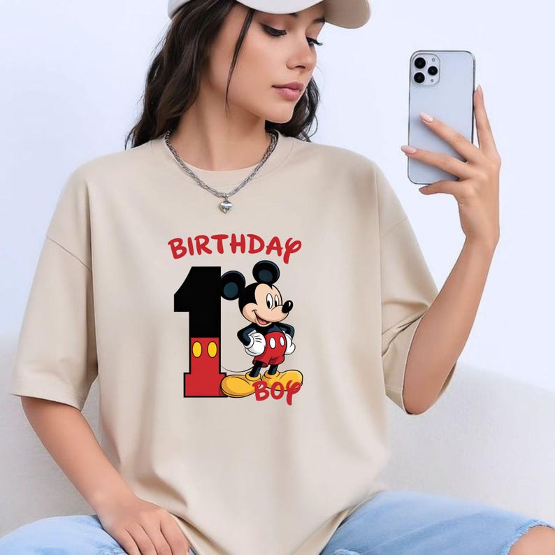 May include: Beige t-shirt with a graphic of Mickey Mouse, the number 1, and the words "BIRTHDAY BOY" in red. Mickey is wearing red shorts, black gloves, and yellow shoes. The shirt is a casual style.