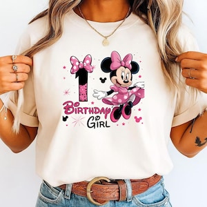 May include: A cream t-shirt with a Minnie Mouse graphic in a pink polka-dot dress and bow. The number 1 and the words "Birthday Girl" are in pink. The design includes stars and Mickey Mouse silhouettes.