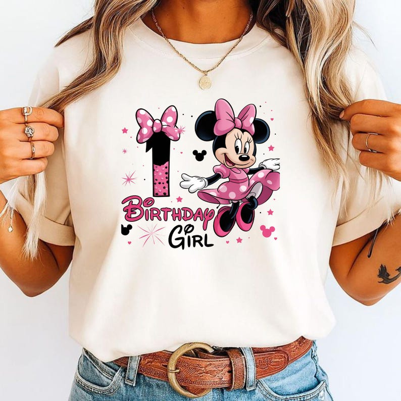 May include: A cream t-shirt with a Minnie Mouse graphic in a pink polka-dot dress and bow. The number 1 and the words "Birthday Girl" are in pink. The design includes stars and Mickey Mouse silhouettes.