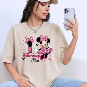 May include: Beige t-shirt with a Minnie Mouse graphic. The design includes a pink bow, dress, and the number 1. The words "Birthday Girl" are in pink. The t-shirt is a relaxed fit.