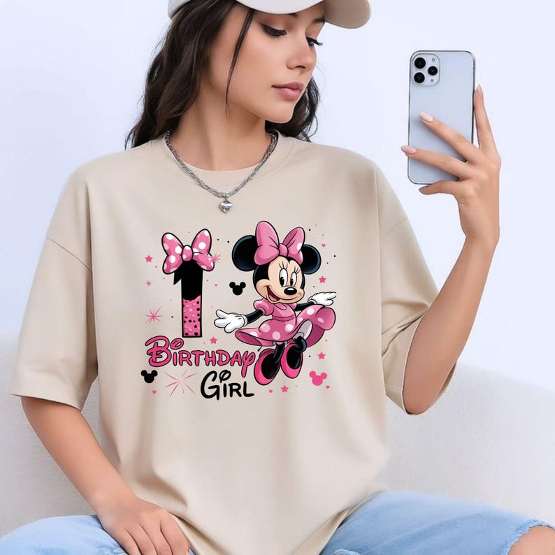 May include: Beige t-shirt with a Minnie Mouse graphic. The design includes a pink bow, dress, and the number 1. The words "Birthday Girl" are in pink. The t-shirt is a relaxed fit.