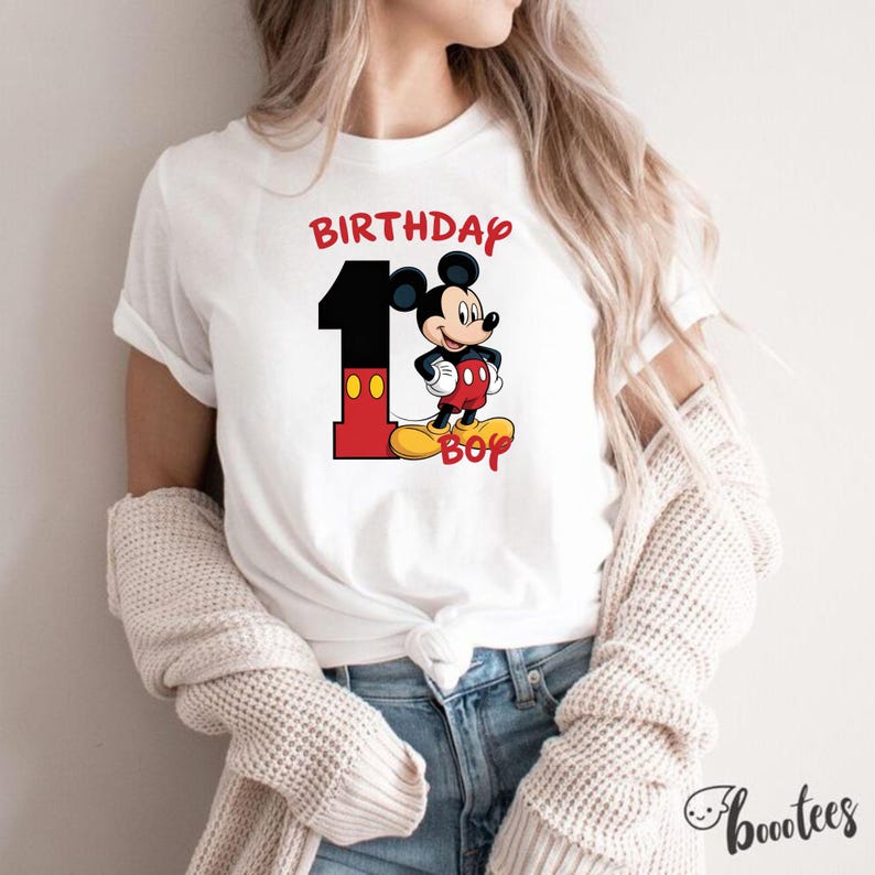 May include: White t-shirt featuring a colorful graphic of Mickey Mouse with the text "BIRTHDAY 1 BOY". The number 1 is black and red, and Mickey Mouse is in his classic outfit. The shirt is a casual style.
