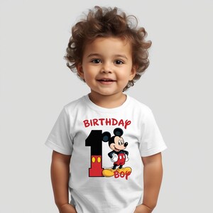 May include: A white t-shirt with a colorful graphic of Mickey Mouse and the number 1. The word "BIRTHDAY" is printed in red above the graphic, and the word "BOY" is printed in yellow below. The shirt is for a one-year-old.